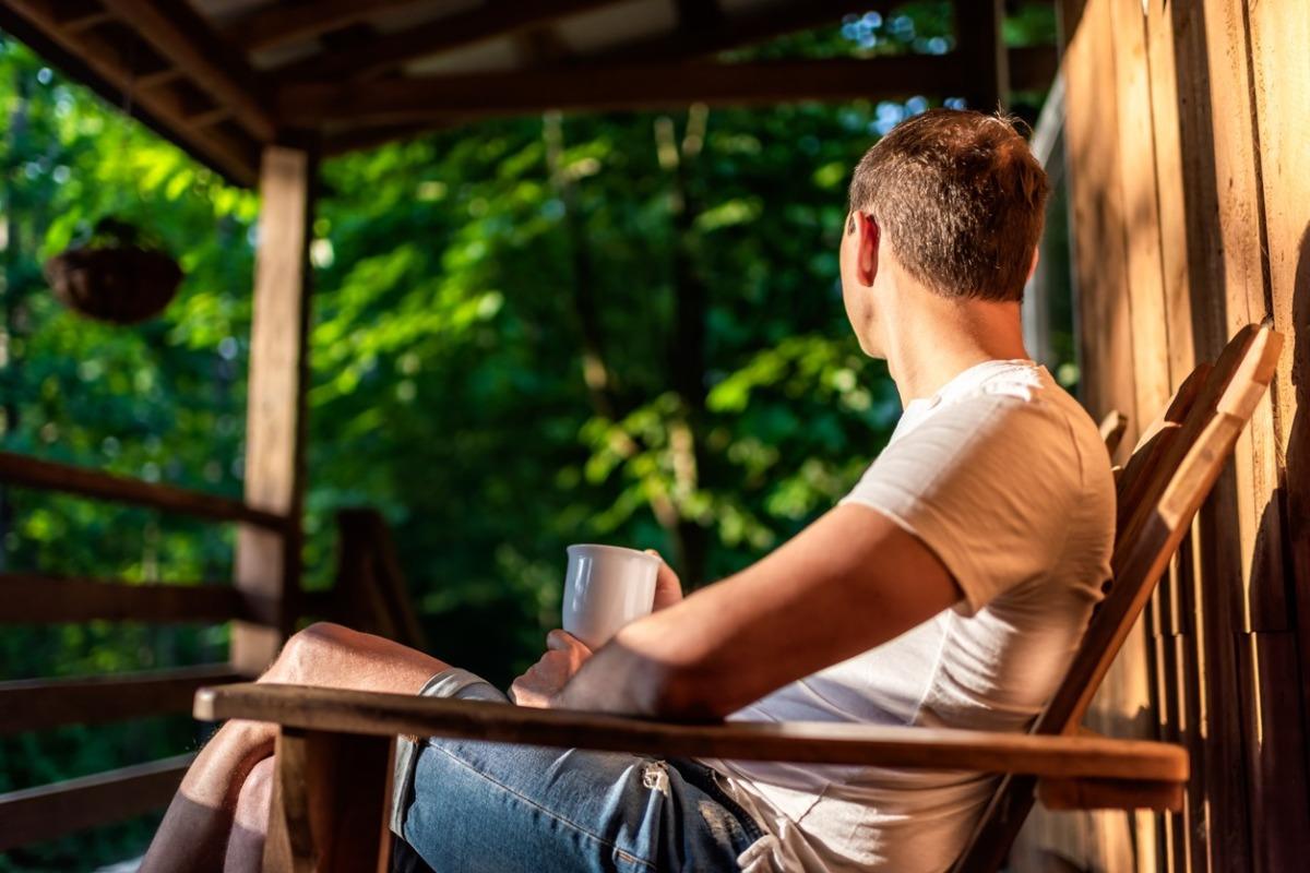 Man sitting on rocking chair on porch of rustic cabin, watching sunrise in morning drinking coffee from cup mug